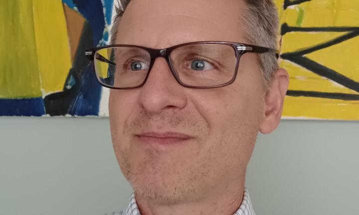 close up of a man's smiling face. he has facial hair stubble and is wearing glasses in front of an abstract painting with mostly yellow and blue