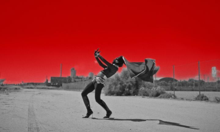 figure in black and high heels leaning back against an industrial ruin and red sky