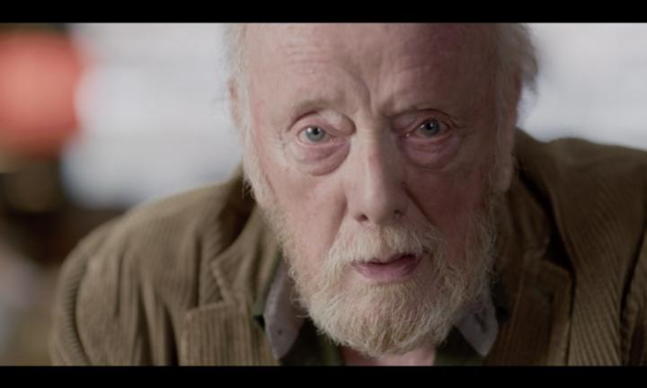 close up of an elderly man with blue green eyes and a white beard and mustache wearing a brown cordouroy jacket staring at the camera