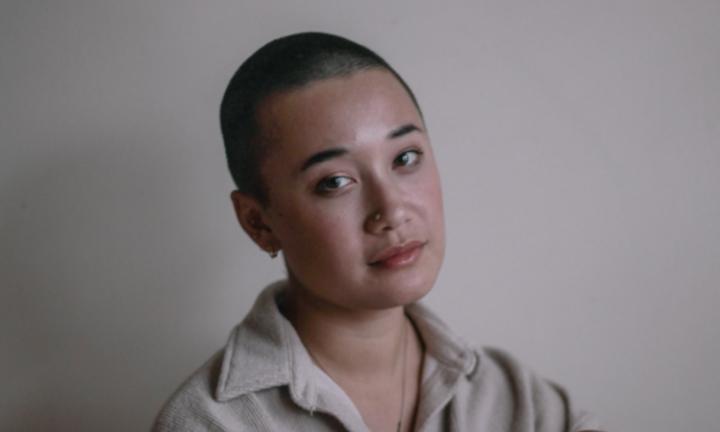 person with shaved head and dark eyes, head tilted toward the camera, in a thick collared tan shirt against a tan background