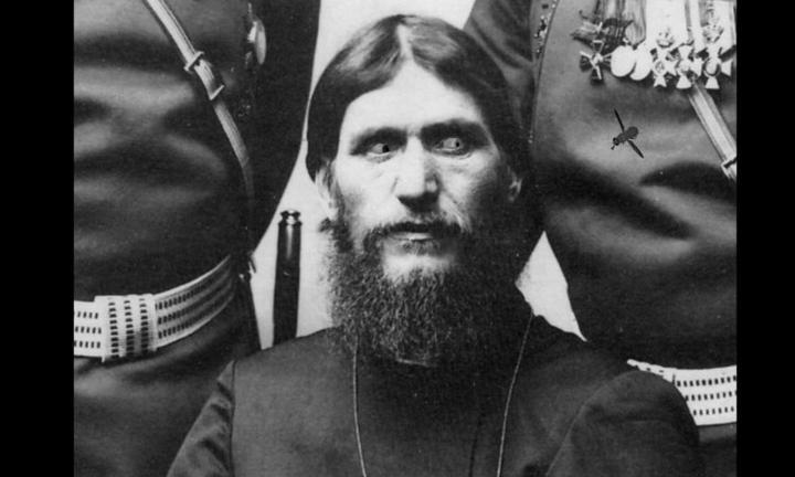 Bearded man seated between two standing figures, all in uniform. Figure on the left has many medals hanging from his chest. Seated figure's eyes are altered to bulge at superimposed fly