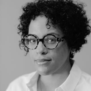 Black and white portrait of author with short to medium curly hair, and round turtleshell glasses.
