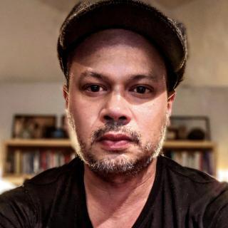 man in a cap with a gray and brown short beard and dark t-shirt with bookcase in the background