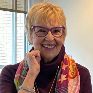 Jane Barnard, women with short blonde hair, wearing purple eyeglasses and a matching purple turtleneck 