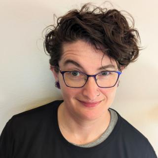 person with short curly brown hair, blue glasses, and a nose ring with eyebrow cocked, smiling at the camera