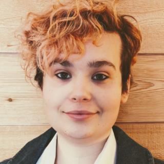 Person with short dark hair and a wavy orange lock, and dark eyes against a wall of rough wood