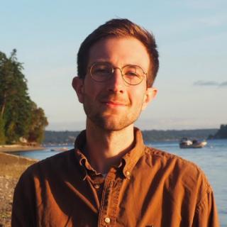 man in glasses wearing a burnt Sienna button-down shirt standing in front of a lake