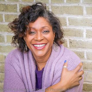 A woman in a lavender sweater smiling in front of a beige brick wall